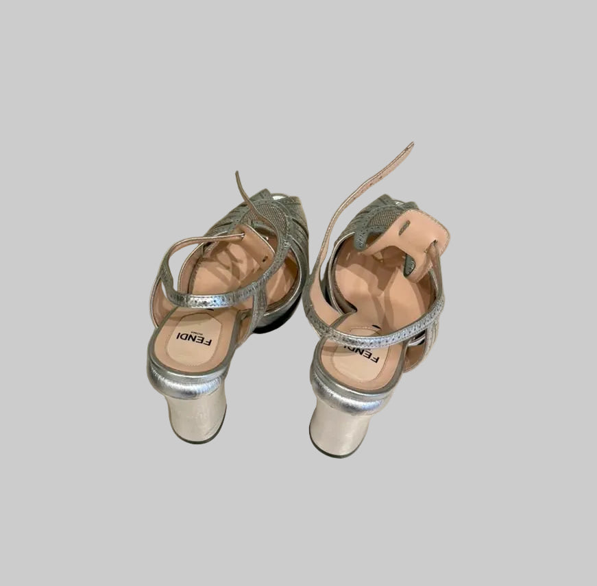 A pair of metallic silver heeled sandals with cutout straps and metallic block heels, shown from a top-down perspective.