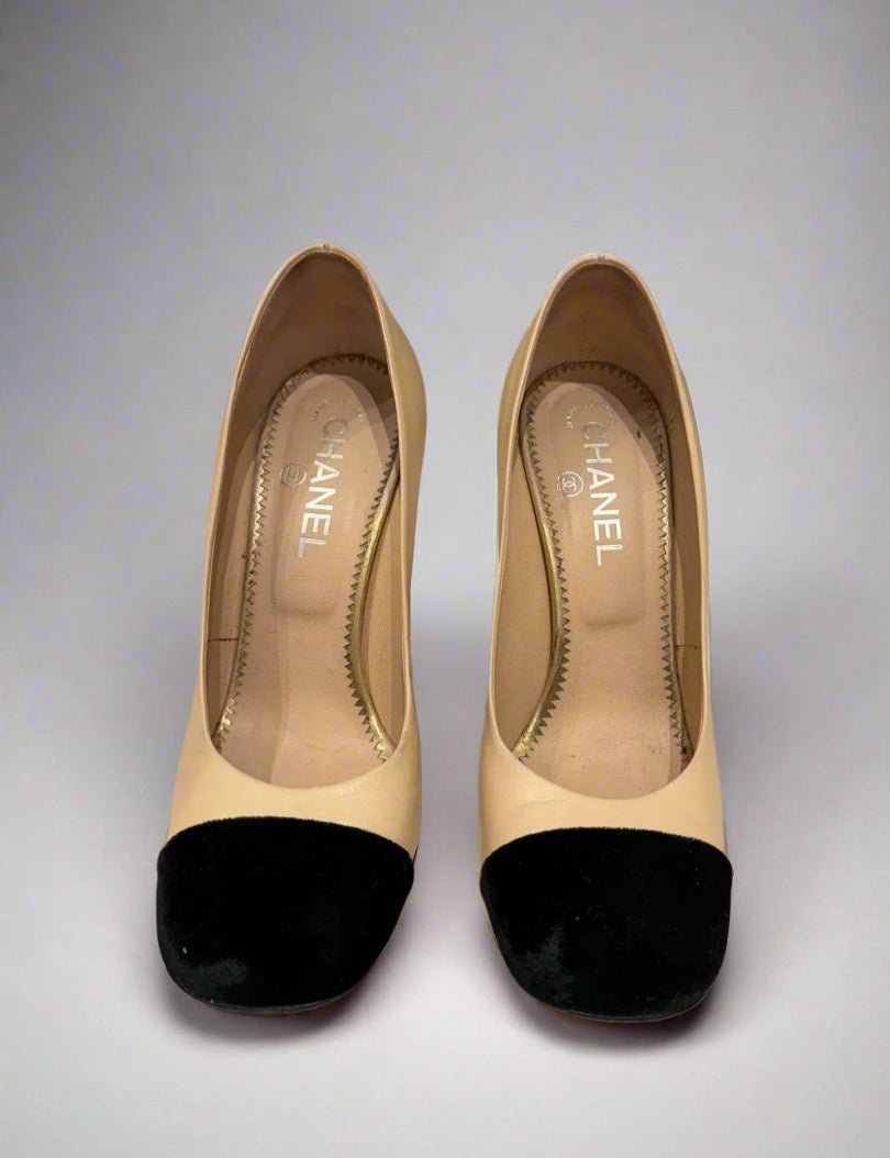 Front view of CHANEL Cap-Toe Two-Tone Pumps in size 38C. The shoes feature a classic cap-toe design with black velvet at the toe and soft beige leather on the rest of the upper. They are displayed against a checkered background