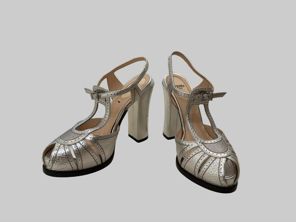 A pair of metallic silver high-heeled slingback sandals with cutout straps and block heels.