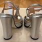 A pair of metallic silver block-heel sandals with T-strap details, shown from the back.