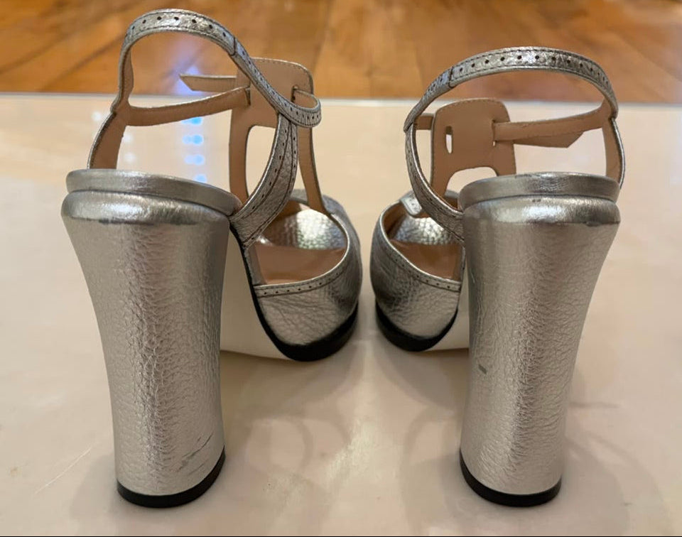 A pair of metallic silver block-heel sandals with T-strap details, shown from the back.