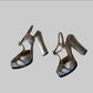 A pair of metallic silver high-heeled sandals with block heels and ankle straps.