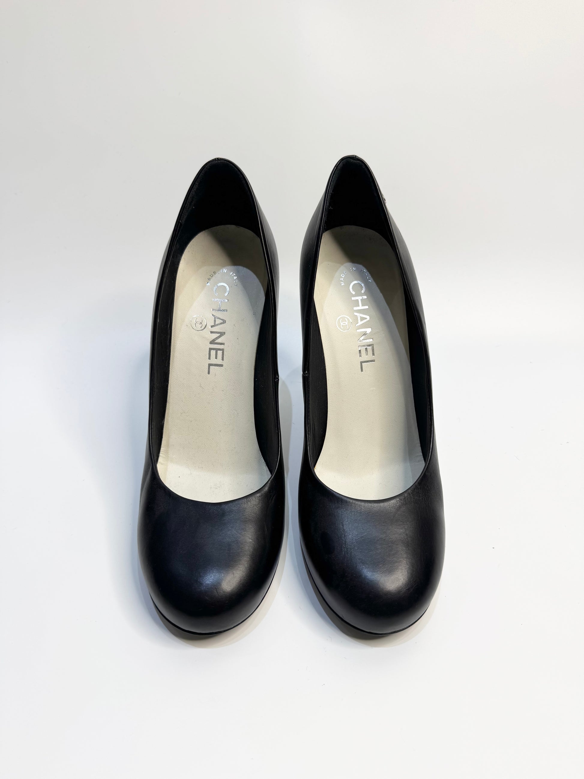 A pair of CHANEL black leather pumps displayed from the front, showcasing their sleek, pointed-toe design.